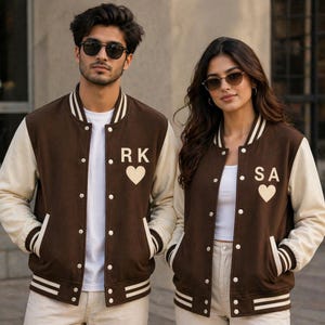 Puede incluir: Dos personas con chaquetas universitarias marrones y crema a juego. Las chaquetas tienen rayas blancas en las mangas y el cuello, con iniciales y un símbolo de corazón en el pecho. Las chaquetas tienen cierre de botones y bolsillos.