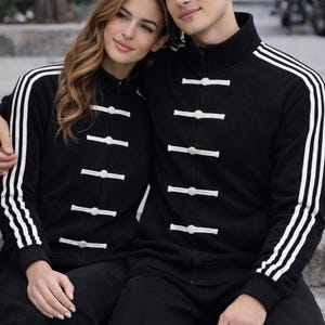 Puede incluir: Conjuntos de chándal negros a juego con rayas y detalles blancos. Las chaquetas tienen un cierre de botón único. La pareja lleva los chándales al aire libre. Los chándales son una opción elegante y cómoda para el uso diario.