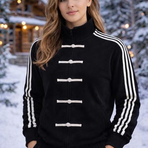 May include: Black zip-up jacket with white stripes on the sleeves and white horizontal closures. The jacket has a high collar and is worn with black pants. The background shows a snowy landscape with a wooden cabin.