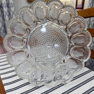 May include: A clear glass egg plate featuring a circular center with a hobnail texture surrounded by twelve oval-shaped indentations, each with a hobnail base and a fluted rim. The plate is supported by a thin, silver-toned metal wire stand.