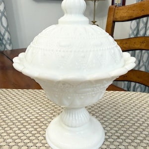 May include: A white milk glass compote dish with a matching lid, featuring a raised hobnail and diamond pattern, a scalloped rim, and a pedestal base. The lid is topped with a rounded finial, and the entire piece has a smooth, opaque finish.