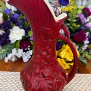 May include: A red ceramic pitcher with a white interior and a handle. The pitcher features a raised leaf and berry design. The pitcher is likely used for decorative purposes or for serving beverages.