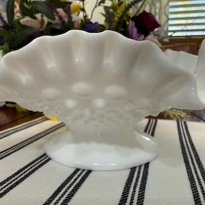 May include: A white, ruffled-edge glass bowl with a pedestal base. The bowl has a textured, hobnail-like pattern on the sides and a scalloped rim. The bowl is sitting on a striped fabric.