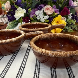 May include: Three brown ceramic soup bowls with handles, featuring a cream-colored rim and vertical ribbed design. The bowls are set on a striped tablecloth, with a floral arrangement in the background.