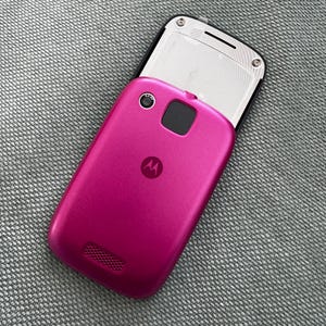 Op de afbeelding: Een magentakleurige Motorola mobiele telefoon met een schuifbare achterkant. Het toestel is voorzien van een cameralens aan de achterzijde, een zwarte vierkante sensor, een rond Motorola-logo en een luidsprekerrooster. Bovenaan is een zilverkleurig paneel.