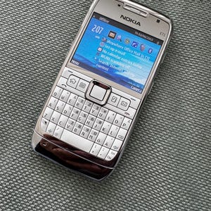 Może przedstawiać: Srebrny telefon komórkowy Nokia E71 z pełną klawiaturą QWERTY i kolorowym wyświetlaczem. Ekran pokazuje godzinę 2:07, datę 03.06.2010 oraz różne ikony systemowe, w tym pocztę e-mail, kalendarz i połączenie Bluetooth.