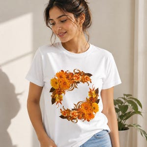 Op de afbeelding: Wit t-shirt met een bloemenkrans ontwerp in oranje, geel en bruin. De krans bestaat uit verschillende bloemen en bladeren, die een cirkelvormig patroon vormen. Het t-shirt is gemaakt van zacht materiaal.