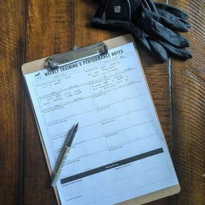 Può includere: Un blocco per appunti con "WEEKLY TRAINING & PERFORMANCE NOTES" stampato sopra, una penna e un paio di guanti neri. Il blocco per appunti ha una clip argentata ed è su una superficie di legno. I guanti sono in pelle.