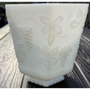 May include: A white glass vase with a square base and a slightly flared top. The vase has an embossed design of grape clusters and leaves. The textured surface adds depth to the design, creating a classic, elegant look. A vintage style piece.