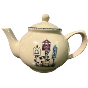 May include: A cream-colored ceramic teapot with a matching lid, featuring a decorative illustration of three birdhouses on a post, a bird in a cage, and a potted plant. The teapot has a curved handle and a spout, with a green speckled rim.