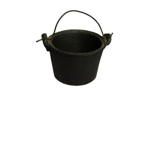 May include: A small, black, cast iron cauldron with a matte finish. The vessel features a wide, open top, a tapered body, and two side lugs that hold a thin, arched metal wire handle that extends above the rim of the container.