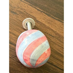 May include: A decorative cabinet knob with a rounded, oval shape. The knob features a pattern of alternating red and light blue diagonal stripes. It has a silver-coloured metal base and screw.