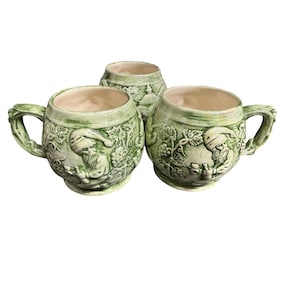 May include: Three ceramic mugs with a light green glazed finish. Each mug features a relief design of a bearded figure wearing a cap, surrounded by grapevines and clusters of grapes. The mugs have a rounded body, a wide rim, and a curved handle.