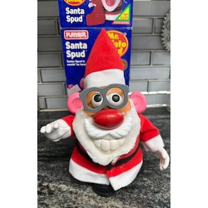 May include: A Santa Spud toy figure wearing a red and white Santa hat, a red coat with white trim, and grey plastic glasses. The figure has a large red nose, a white mustache, and prominent white teeth, positioned in front of its original box.