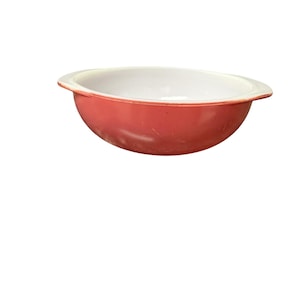 May include: A round, shallow bowl with a smooth, coral-red exterior and a contrasting white interior. The bowl features two small, integrated handles on opposite sides, providing a simple, functional design for serving or food preparation.