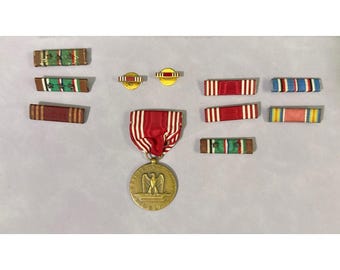 May include: A collection of military medals and ribbons. The central medal is gold-toned with an eagle emblem. Ribbons feature various color combinations, including red, white, blue, green, and yellow. The items are arranged on a light gray surface.