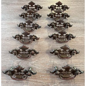 May include: A set of ten matching dark bronze-toned metal drawer pulls arranged in two vertical columns. Each pull features a decorative backplate with a central cutout and a curved, swinging handle attached to two mounting posts on the back.