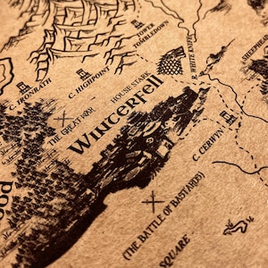 May include: A detailed map with a vintage aesthetic, featuring intricate illustrations of landscapes, cities, and geographical features. The map is printed on aged paper, with the word "Winterfell" prominently displayed. The map includes text such as "The Great War" and "The Battle of Bastards".