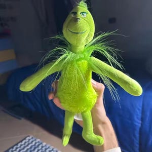May include: A plush green character with a smiling face, long arms, and legs. The character has a tuft of hair on top of its head and a fluffy collar. The character is holding its arms out to the sides.