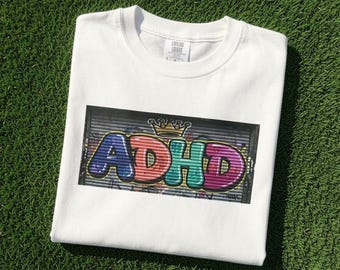 May include: A white short-sleeve t-shirt featuring a rectangular graphic print on the chest. The graphic displays the text ADHD in colourful, bubble-style graffiti lettering with a gold crown icon above it, set against a dark metal shutter background.