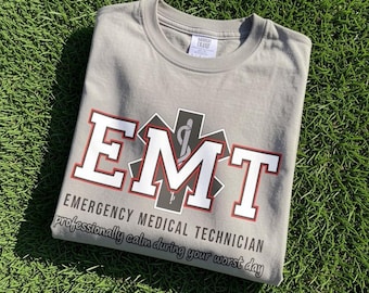 May include: A light grey crew neck t-shirt featuring the white text EMT with a red outline and a black Star of Life symbol. Below, the text reads EMERGENCY MEDICAL TECHNICIAN and professionally calm during your worst day in black lettering.