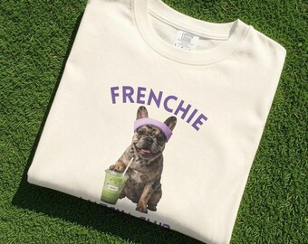 May include: A cream-coloured crewneck sweatshirt featuring a graphic of a French Bulldog wearing a light purple headband and holding a green iced beverage. The text "FRENCHIE" is printed in purple above the dog illustration on the front.