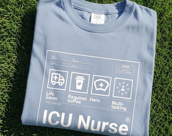 May include: A light blue t-shirt featuring a white graphic print that resembles a form. The design includes icons for an ambulance, coffee cup, nurse cap, and multitasking, with the text "Life Saver," "Requires coffee," "Hero," and "ICU Nurse."