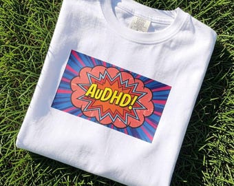 May include: A white short-sleeved t-shirt featuring a rectangular graphic print on the chest. The graphic displays the text AuDHD! in yellow letters inside a red and orange comic book style speech bubble, set against a blue and purple sunburst background.
