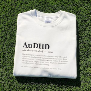 Könnte beinhalten: Ein weißes T-Shirt mit Rundhalsausschnitt und schwarzem Text, der AuDHD lautet, gefolgt von einer phonetischen Aussprache, der Klassifizierung als Substantiv und einem beschreibenden Absatz über die neurologische Erfahrung.