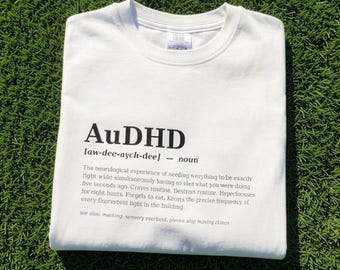 May include: A white crewneck t-shirt featuring black text that reads AuDHD, followed by a phonetic pronunciation, the noun classification, and a descriptive paragraph detailing the neurological experience of needing everything to be exactly right.