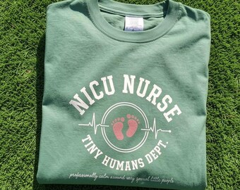 May include: A sage green crew neck t-shirt featuring white text that reads NICU NURSE TINY HUMANS DEPT. with a central graphic of pink baby footprints inside a circle intersected by a heartbeat line and smaller text below reading professionally calm around very special little people.