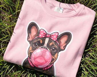 May include: A light pink crewneck sweatshirt featuring a graphic print of a French Bulldog with a pink polka dot bow on its head and a pink bubble gum bubble in its mouth. The dog wears a pink collar and has large, dark, expressive eyes.