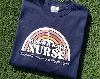 May include: A navy blue t-shirt featuring a graphic of a multi-coloured rainbow with a small crescent moon. The text reads "MOTHER BABY NURSE" in large white letters, followed by the script text "two patients. one room. zero sleep for anyone."