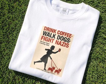 May include: A white t-shirt featuring a graphic print of a silhouette figure holding a coffee cup and walking two dogs on leashes. The text reads: DRINK COFFEE, WALK DOGS, FIGHT NAZIS, and She never misses a morning routine, with smaller text below.