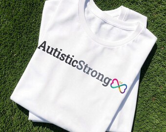 May include: A white short-sleeved t-shirt featuring the black text AutisticStrong printed across the chest, followed by a small, colourful infinity symbol with a gradient of blue, green, yellow, orange, and purple hues on the right side.