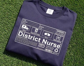 May include: A navy blue t-shirt featuring a white graphic print designed like a shipping label. The text reads District Nurse, 100% ORGANIC, and includes icons for an ambulance, coffee cup, nurse cap, and multi-tasking symbol with various chemical elements.