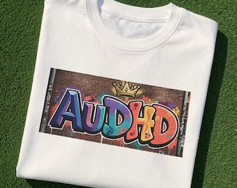 May include: A white crew neck t-shirt featuring a rectangular graphic print on the chest. The graphic displays the text AUDHD in colourful graffiti-style lettering against a brick wall background, topped with a small yellow crown illustration.