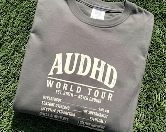 May include: A charcoal grey crew neck t-shirt featuring white text that reads AUDHD WORLD TOUR EST. BIRTH - NEVER ENDING, followed by a list of terms including HYPERFOCUS, SENSORY OVERLOAD, EXECUTIVE DYSFUNCTION, 3:00 AM, and THE SUPERMARKET.