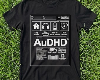 May include: A black short-sleeved t-shirt featuring a white graphic print that resembles a shipping label. The design includes icons for a house, headphones, a battery, and a heart, with the text AuDHD, 100% ORGANIC, and various ingredient lists.