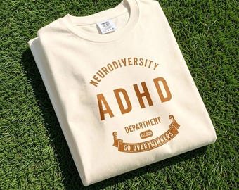May include: A cream-coloured crewneck t-shirt featuring brown text that reads NEURODIVERSITY ADHD DEPARTMENT EST. 2026 GO OVERTHINKERS. The design includes an arched header and a ribbon graphic at the bottom, with a Comfort Colors brand label visible.