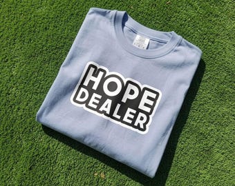 May include: A light blue crew neck t-shirt featuring a black rectangular graphic with a white border and the text HOPE DEALER in bold white capital letters. The shirt has a visible interior neck label and is folded flat on a textured surface.