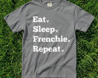 May include: A grey t-shirt featuring the white text "Eat, Sleep, Frenchie, Repeat", with a small dog illustration beside each word. The text is stacked. The t-shirt is made of a soft material and is a casual wear item.