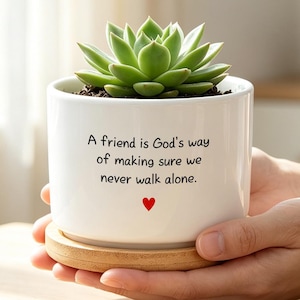 May include: A white ceramic cylindrical planter containing a green succulent plant. The planter features the black text: A friend is God's way of making sure we never walk alone, with a small red heart icon below the text, resting on a wooden base.