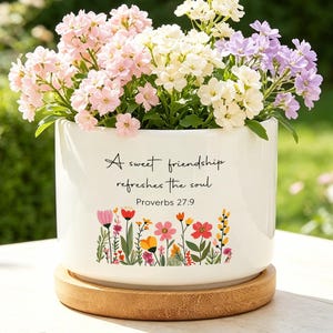 Peut inclure: Un pot de fleurs cylindrique en céramique blanche avec une soucoupe en bambou, orné d'un motif floral coloré et du texte noir "A sweet friendship refreshes the soul Proverbs 27:9", contenant un bouquet de fleurs roses, blanches et lilas.