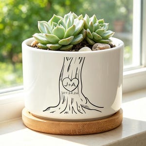 May include: A white ceramic cylindrical planter with a black line drawing of a tree trunk featuring a heart carved with the letters J+A and the date 07.14.26. The planter sits on a round bamboo saucer and contains a green succulent plant.