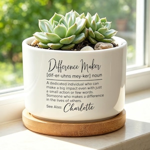 May include: A white ceramic cylindrical planter with a bamboo saucer containing a green succulent. The planter features black printed text: Difference Maker [dif-er-uhns mey-ker] noun, a definition, and the name Charlotte.