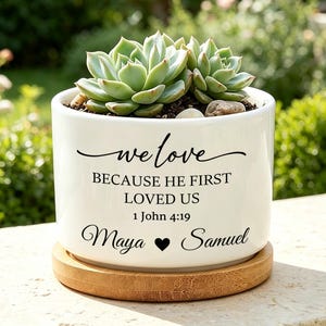 Peut inclure: Un pot de fleurs cylindrique en céramique blanche avec une soucoupe en bambou contenant une plante succulente verte. Le pot porte l'inscription noire : we love BECAUSE HE FIRST LOVED US 1 John 4:19 Maya cœur Samuel sur la face avant.