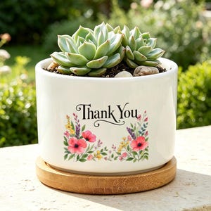 Op de afbeelding: Een cilindervormige witte keramische plantenpot met een bamboe schoteltje. De voorzijde is voorzien van de zwarte tekst Thank You boven een kleurrijk aquarel bloemstuk met rode klaprozen, gele wilde bloemen en groen blad.