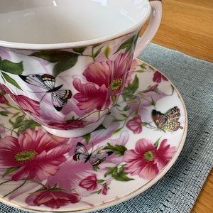 Peut inclure: Une tasse à thé et sa soucoupe en porcelaine blanche ornées d'un motif floral vibrant composé de grandes fleurs roses, de feuilles vertes et de plusieurs papillons aux ailes détaillées. Le bord de la tasse et de la soucoupe est doré.