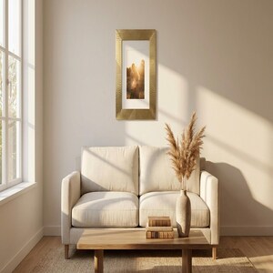 May include: A gold rectangular picture frame with a textured pattern hangs on a cream wall. The frame surrounds a photograph with a white mount. Below, a cream sofa and wooden coffee table are visible.